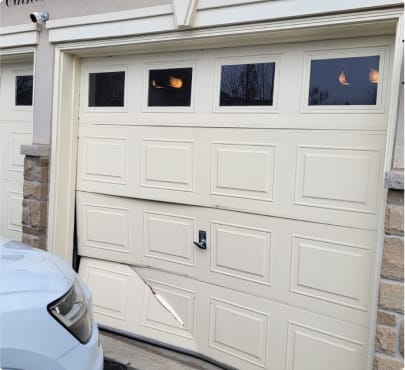 A garage door with a dent at the bottom right side, with a white car parked partially in view on the left, recently fixed by our locksmith based on an appreciated client testimonial.