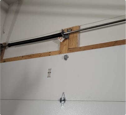 A garage interior wall corner showing a metal torsion spring mechanism for a garage door, mounted on wooden blocks with a steel horizontal support bar, recently serviced by a locksmith according to a satisfied client's testimon