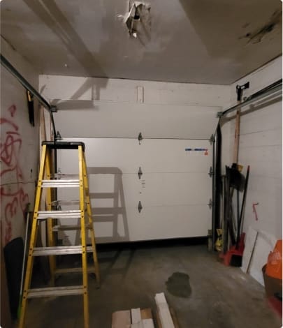 A cluttered and unfinished garage interior with a white door, testimonial from a satisfied client framed against the wall beside a yellow ladder, various tools and items stored at the sides, and a visible water