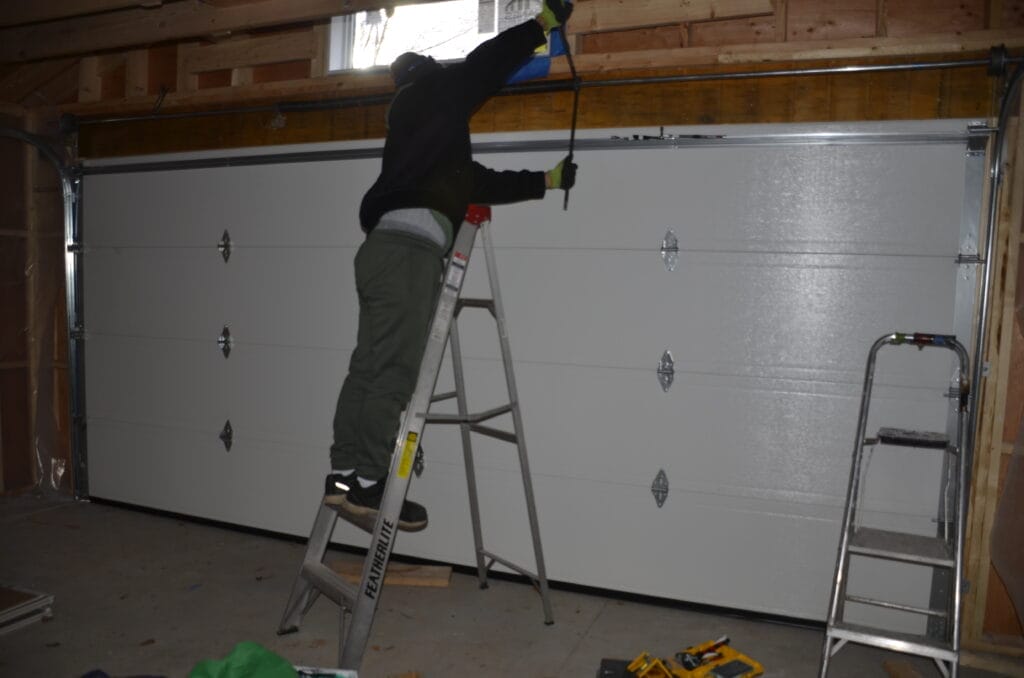 A man is standing on a ladder while making some home repairs specifically replacing a garage door in Oakville, with tools scattered on the ground below.