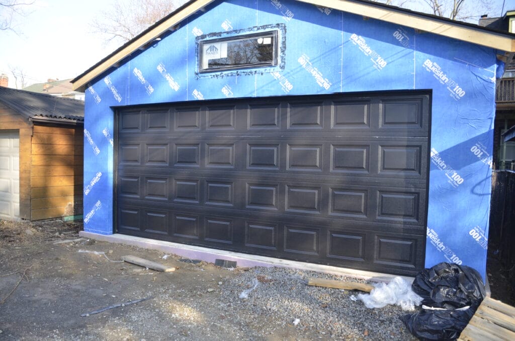 A residential garage under construction, featuring a newly installed black sectional garage door from Icarus Services, is set against a building wrapped in blue weather-resistant sheathing material.