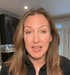 A female customer with long, light brown hair is indoors, standing in a kitchen. she is wearing a black zip-up jacket and appear to be speaking or mid-sentence about home repairs in Toronto provided by Icarus services . Cabinets, a countertop, and a plant are visible in the background.