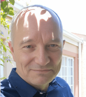 A middle-aged male client with a receding hairline, wearing a navy blue shirt, smiles slightly while standing outdoors in front of a building with a white window. Dappled sunlight and some leaves are visible in the top left corner of the image, capturing a serene moment amidst home repairs .