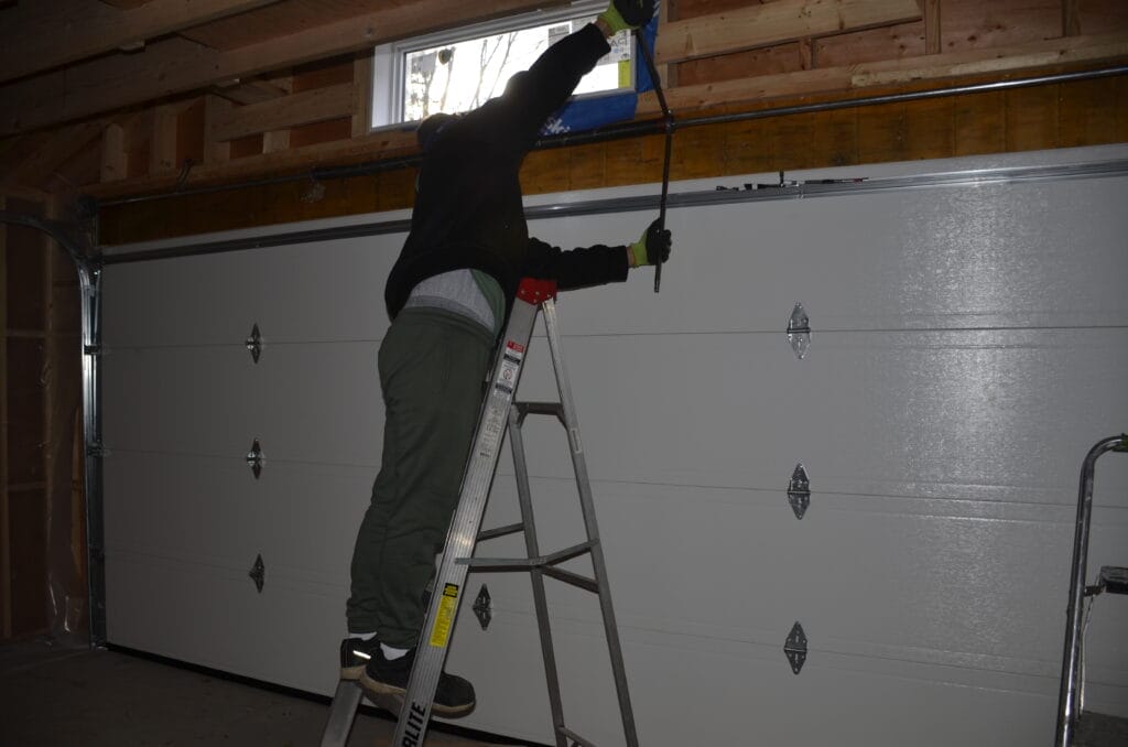 A male technician is standing on a ladder while making some home repairs specifically replacing a garage door in Oakville, with tools scattered on the ground below.