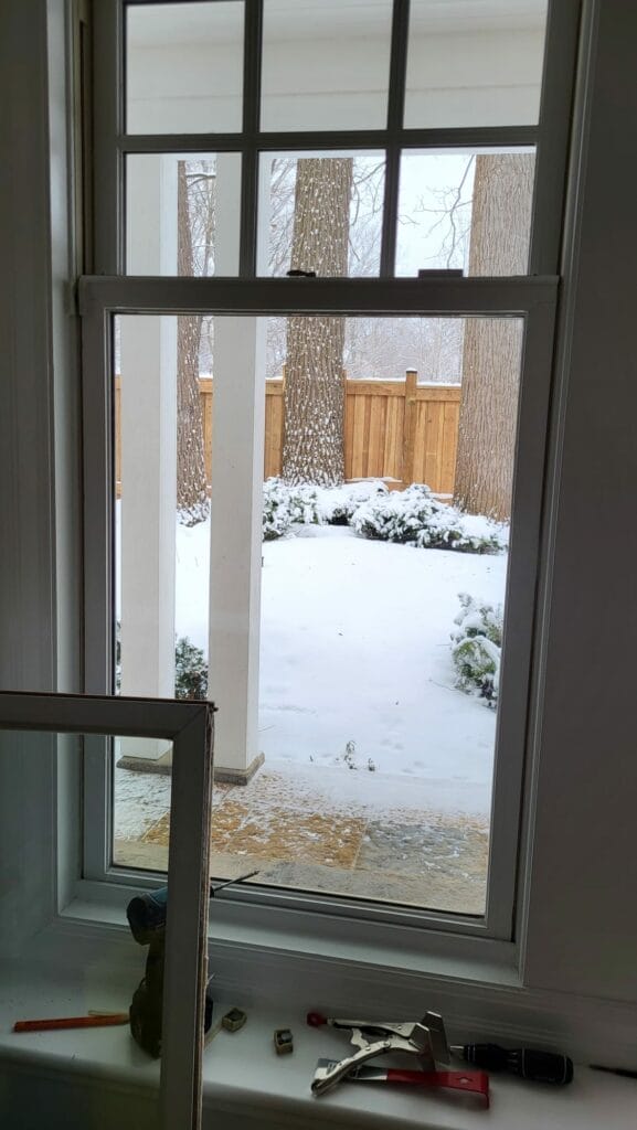 A view through a window during winter in Toronto showing a snowy backyard with several trees and a wooden fence. Snow covers the ground and bushes. Inside, there's a detached window screen, a few tools, and other small objects on the windowsill.