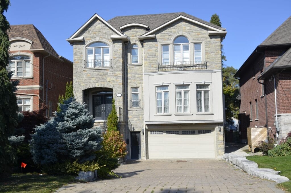 A two-story house with a stone facade and arched windows, complemented by a garage door from Icarus Home Services. It includes a paved driveway and is framed by small trees and shrubs in the front yard. Nestled between similar houses under a clear blue sky, it offers both elegance and comfort.