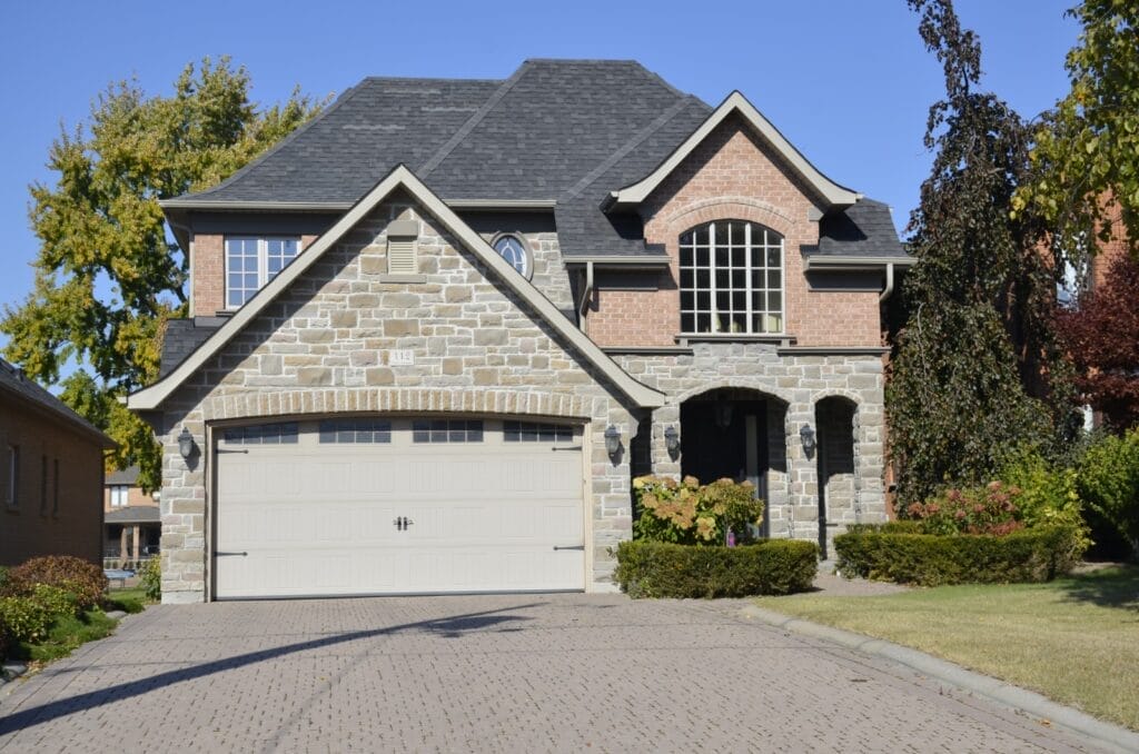 A two-story house with a gray stone facade, brick accents, and arched windows stands proudly. Under the care of Icarus Home Services, the property features a double garage with a light-colored door and a paved driveway. Neatly trimmed shrubs and trees surround it under a clear blue sky.