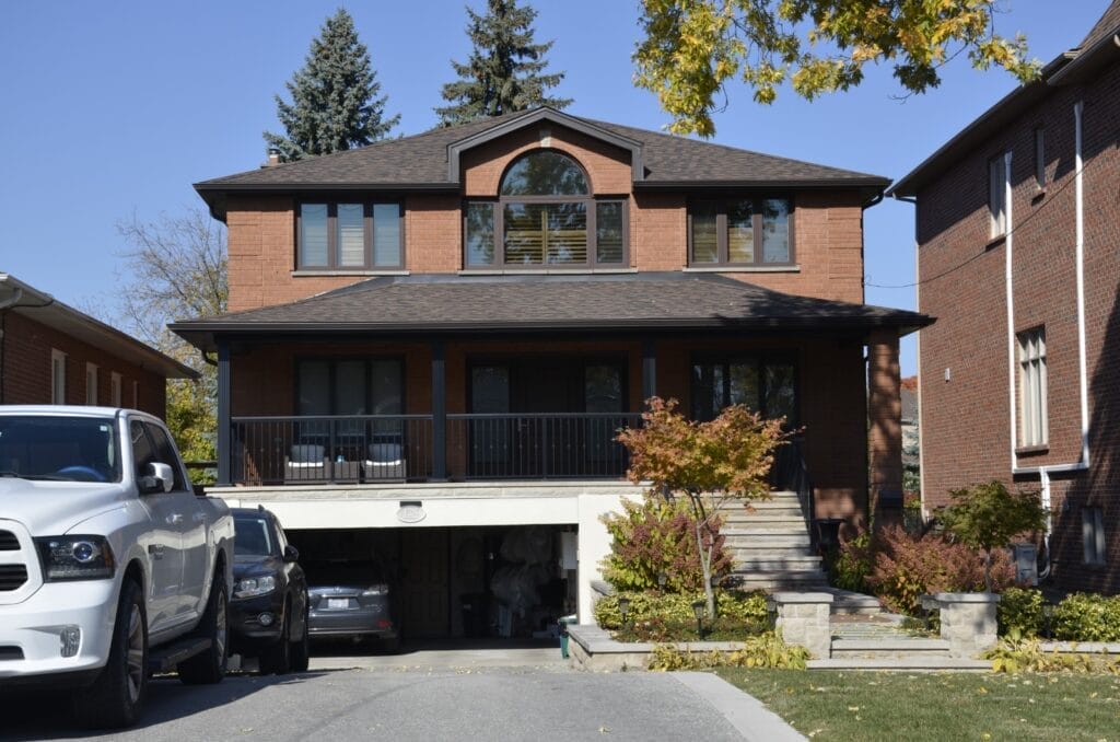 A two-story brick house with a large arched window on the top floor. It features a covered front porch with chairs and a driveway leading to a garage door maintained by Icarus Home Services. Several cars are parked outside amid autumn trees and shrubs in the front yard.